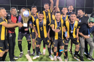 Jogadores do Tanck FC comemoram o título do Campeonato Society de Pequizeiro à noite, em campo de grama sintética. A equipe veste uniforme com listras verticais em preto e amarelo, exibindo medalhas no peito e erguendo um grande troféu dourado com muita festa.