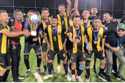 Jogadores do Tanck FC comemoram o título do Campeonato Society de Pequizeiro à noite, em campo de grama sintética. A equipe veste uniforme com listras verticais em preto e amarelo, exibindo medalhas no peito e erguendo um grande troféu dourado com muita festa.