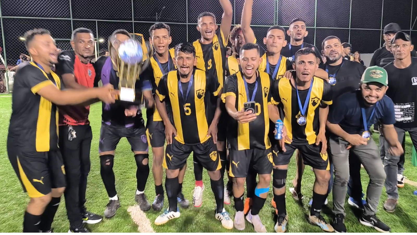 Jogadores do Tanck FC comemoram o título do Campeonato Society de Pequizeiro à noite, em campo de grama sintética. A equipe veste uniforme com listras verticais em preto e amarelo, exibindo medalhas no peito e erguendo um grande troféu dourado com muita festa.