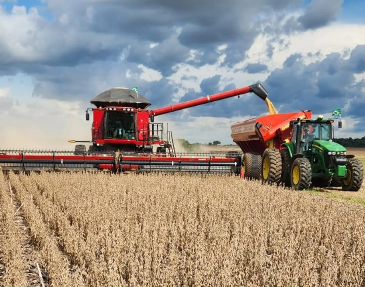 Colheitadeiras em campos de soja no Tocantins, com estradas rurais precárias e caminhões em fila ao fundo, ilustrando a discrepância entre produção e logística.