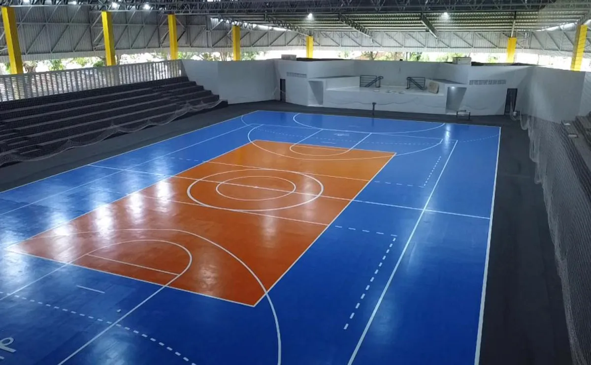 Jogadores de futsal em ação em quadra reformada de ginásio, com torcida ao fundo e placar eletrônico