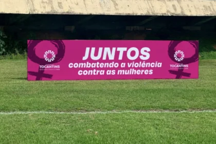 Jogadores de futebol do Campeonato Tocantinense em campo com camisetas estampadas com a frase 'Juntos combatendo a violência contra as mulheres' durante uma partida.