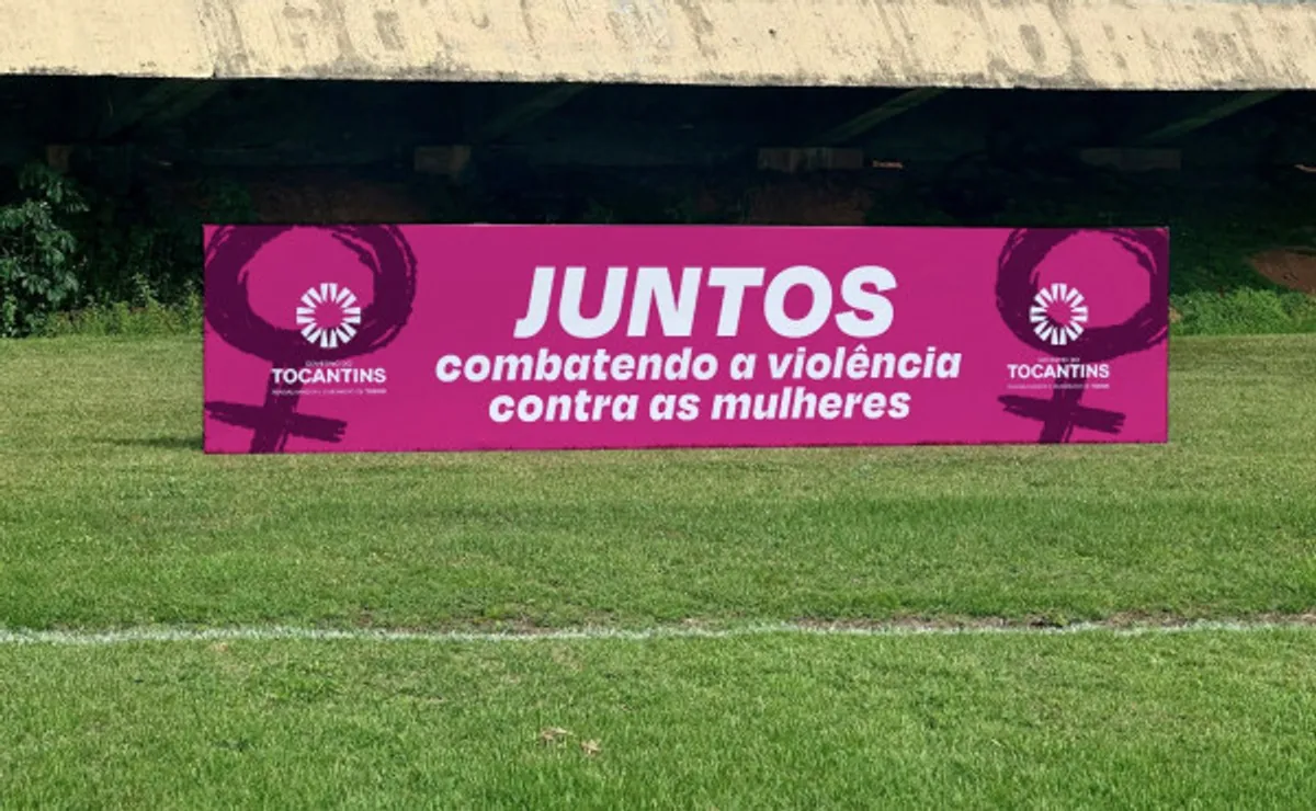 Jogadores de futebol do Campeonato Tocantinense em campo com camisetas estampadas com a frase 'Juntos combatendo a violência contra as mulheres' durante uma partida.
