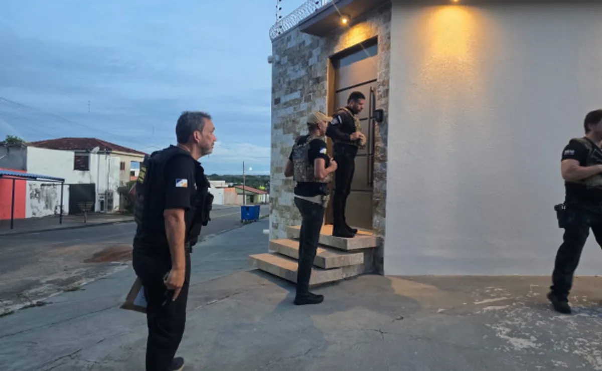 Policiais civis cumprindo mandados de busca e apreensão em uma residência, com documentos e celulares sendo recolhidos como prova em uma operação contra lavagem de dinheiro.