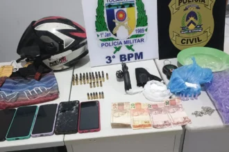 Fotografia de uma mesa branca exibindo diversos materiais apreendidos. Ao fundo, há duas placas com os brasões da Polícia Militar do Tocantins (3º BPM) e da Polícia Civil. Sobre a mesa estão dispostos: um capacete de motociclista sobre uma sacola de tecido estampada, dezenas de munições, um revólver preto com o tambor aberto, um coldre, uma balança de precisão, embalagens plásticas contendo pó branco, um prato verde, um saco grande cheio de pequenos pinos roxos vazios, cinco aparelhos celulares enfileirados, notas de dinheiro em espécie (Reais) e uma corrente prateada.