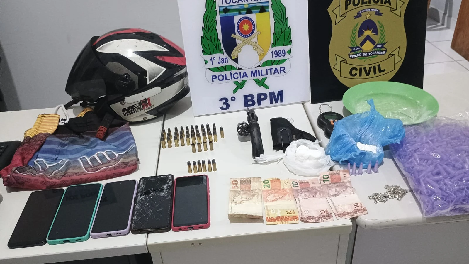 Fotografia de uma mesa branca exibindo diversos materiais apreendidos. Ao fundo, há duas placas com os brasões da Polícia Militar do Tocantins (3º BPM) e da Polícia Civil. Sobre a mesa estão dispostos: um capacete de motociclista sobre uma sacola de tecido estampada, dezenas de munições, um revólver preto com o tambor aberto, um coldre, uma balança de precisão, embalagens plásticas contendo pó branco, um prato verde, um saco grande cheio de pequenos pinos roxos vazios, cinco aparelhos celulares enfileirados, notas de dinheiro em espécie (Reais) e uma corrente prateada.